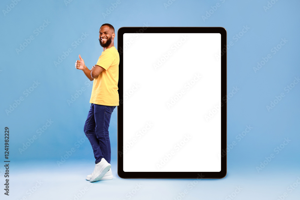 Poster positive african american guy showing thumb up gesture next to huge digital tablet on blue studio ba
