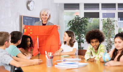 Mature primary school teacher working in a high school tells pupils the history of China in class and holds the national flag .of the country in her hands