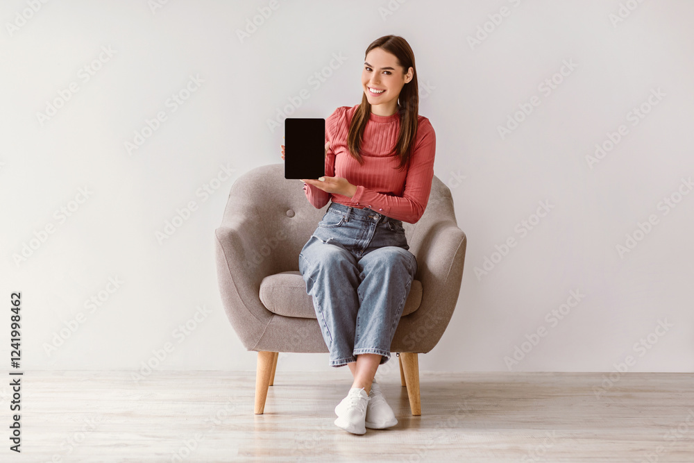 Poster smiling woman in casual wear showing tablet with empty screen, sitting in armchair against white wal