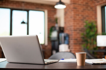 Empty brick wall workspace having table holding laptop, coffee cup and company documents, ready for busy workday. Startup office with no people is well prepared for business meetings and research.