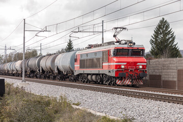 Red electric locomotive is hauling a freight train consisting of tank wagons or tankers with fuel.