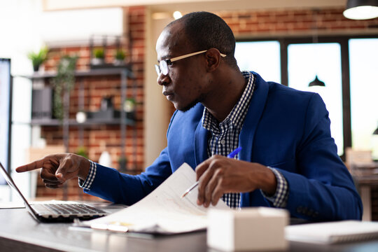 Focused businessman pointing to device screen and reviewing market analysis documents in startup office. Male analyst sitting at desk with laptop and company paperwork, preparing project presentation.