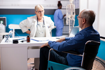 Obraz premium Elderly man in wheelchair sits across clinic desk from friendly senior doctor wearing stethoscope, discussing his treatment options. Male pensioner patient listens carefully to physician at hospital.