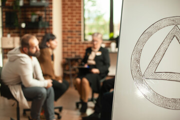 Closeup of aa meeting symbol on white board with ongoing therapy session in background. People engaging in discussions about alcohol addiction with guidance from retired therapist in brick wall office