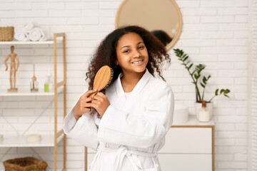 Cute happy African-American girl brushing her hair in bathroom