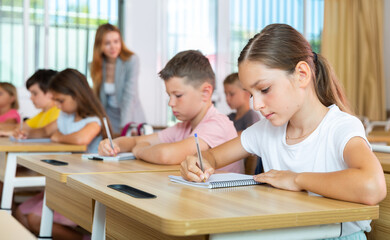 Portrait of concentrated tween girl studying in classroom, listening to schoolteacher and writing in notebook