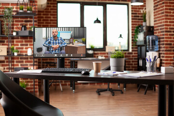 Desktop monitor on wooden table in well arranged room displays virtual meeting with male wheelchair user. Spacious room organized with computers and office supplies for effective planning.