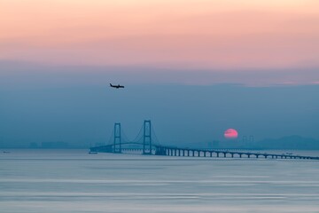 Sunset over a bridge with an airplane flying in the sky near the water