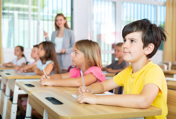 Fototapeta premium Schoolchildren sitting at desks in classroom. Female teacher staning beside desks.