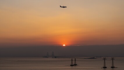 Sunset view with airplane flying over water and bridge in the background