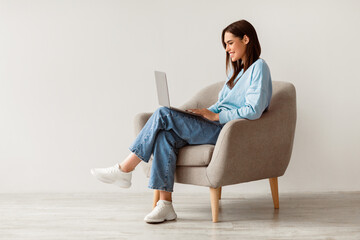 Smiling young woman in casual wear working online, sitting in armchair and using laptop against...