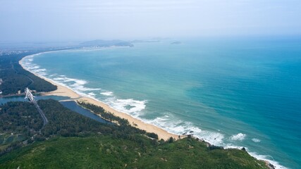 Coastal aerial view showing waves crashing on sandy beach with green hills and a river in the background