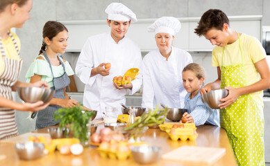 Two experienced chefs, friendly woman and man in white uniform giving culinary lesson to group of inquisitive tweens, teaching basic cooking skills