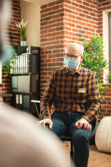 Portrait of senior man seated in brick wall office, attending mental health recovery program. Caucasian pensioner patient with face mask, speaking about his problems during group therapy session.