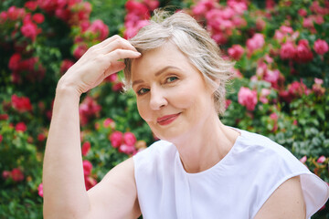 Outdoor summer portrait of mature woman with grey hair, posing in garden with pink roses