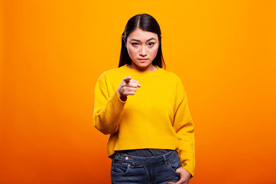 Serious woman in a yellow sweater pointing directly at the camera on an orange background. Young asian lady standing with a stern look and gesturing her index finger towards the viewer.