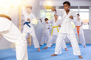 Concentrated black teenager in kimono practicing punches in gym during group martial arts workout....
