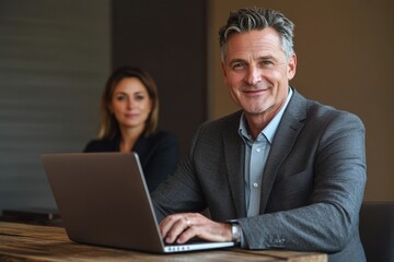 Fototapeta premium Confident businessman working on laptop with colleague in background, showcasing success and collaboration.