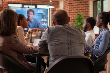 Rearview of entrepreneurs engaged in virtual meeting, brainstorming strategy ideas and using monitor for remote communication. Colleagues sitting at table and listening to client in video call on