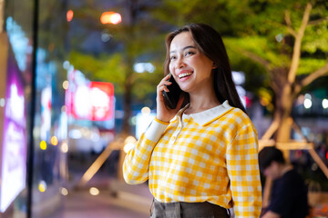 A woman in a yellow checkered  smiles while talking on her phone at night, surrounded by vibrant city lights.