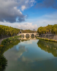 Fototapeta premium Reflection of a historic arched bridge crossing the Tiber River with Vatican City in the background. Ponte Umberto I