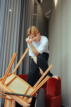 Young man poses defiantly on red chair with tipped furniture