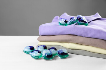 Laundry detergent capsules and clean clothes on white wooden table, closeup. Space for text