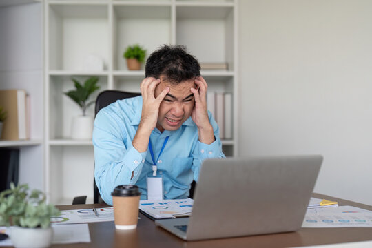 Overwhelmed Asian businessman with head in hands at his desk, dealing with stress while looking at his laptop in a modern office environment.
