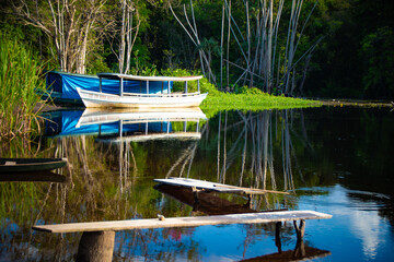 Small wooden boat anchored on a calm river with a larger white boat covered with a blue tarp in the background, surrounded by dense vegetation reflected in the water.