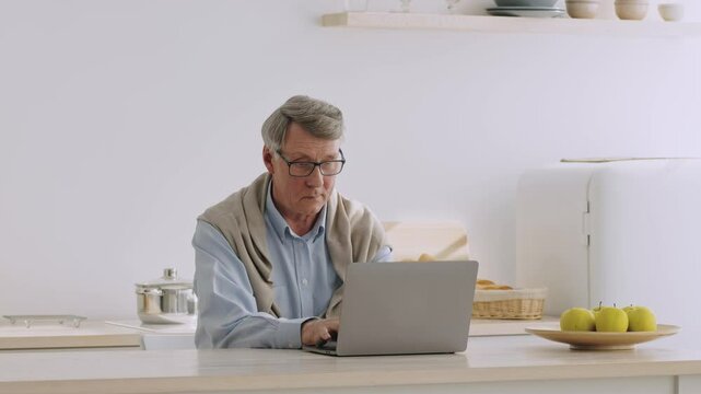 An elderly man in his 60s sits at a kitchen counter using a laptop. He enjoys his retirement lifestyle, surrounded by fresh apples and a basket of bread.
