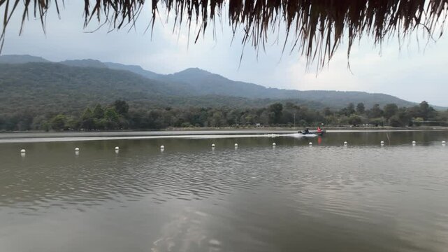 Speedboat passing by a tranquil lake, ripples, viewed from a rustic shelter with wooden framing and thatched roof, against backdrop of forested hills and overcast sky - Huai Tueng Thao Lake Thailand