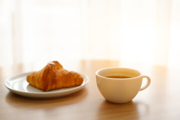 A cup of hot latte and a croissant on a table under soft sunlight.
