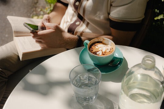 Individual enjoys coffee while reading a book and using a smartphone 