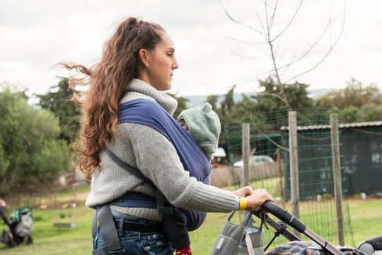 Mother carrying baby in sling while pushing stroller in park