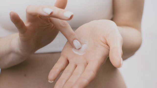 A young woman applies a small amount of moisturizer onto her palm, focusing on her skin care routine. She is in a tranquil environment, enhancing her pampering experience.