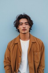 Photo of a young, attractive Japanese man. He stands in front of a light blue wall, wearing a white shirt underneath a brown jacket, looking at the camera and smiling slightly.