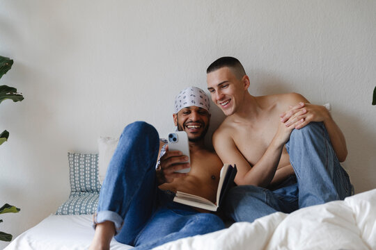 Two shirtless men smiling and relaxing on a bed with a smartphon