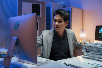 Fototapeta premium Asian businessman working late at night in office looking worried at computer screen with pile of paperwork
