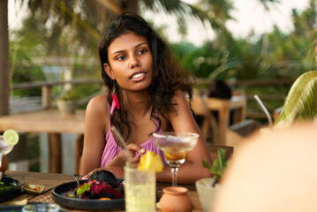 Diverse women enjoy dinner in tropical restaurant. Friends chat over food, drinks. Relaxed atmosphere, palm decor. Girls night out with laughter, cocktails, exotic location.