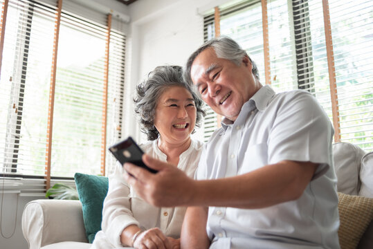 Asian senior couple smiles together on a sofa, sharing a joyful moment as they look at a smartphone, creating warm memories in their retirement. - Powered by Adobe