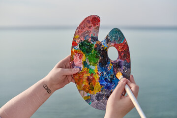 Young female holds colorful artist palette against tranquil seascape, symbolizing creativity and...