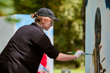 Young female artist with cap and gloves painting on large canvas in sunlight. Surrounded by greenery, she focuses on her work with determination and skill