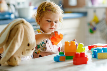 Fototapeta premium Adorable toddler girl with favorite plush bunny playing with educational toys in nursery. Happy healthy child having fun with colorful different plastic blocks at home. Cute baby learning creating.