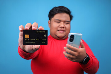 Cheerful Young Asian Fat Man Using Smartphone While Showing Credit Card Isolated on Blue Background