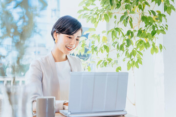 カフェの明るい窓際でパソコンで仕事をする30代の笑顔の女性