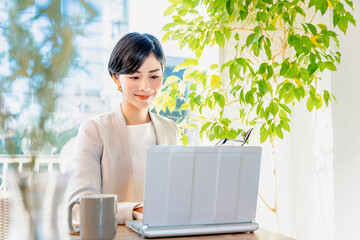 カフェの明るい窓際でパソコンで仕事をする30代の笑顔の女性