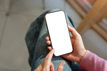 A close-up top view of a woman in jeans sitting indoors and using her smartphone.