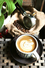 beautifully crafted latte with latte art in a ceramic cup, placed on a saucer, alongside coffee beans spilling from a portafilter onto a burlap sack
