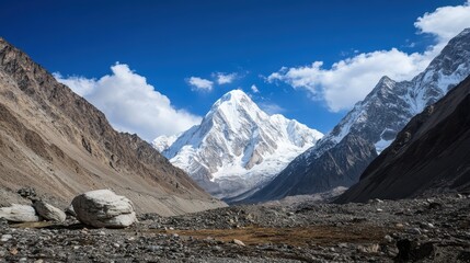 A majestic mountain, adorned with a stunning layer of snow on its peaks, towers magnificently over a tranquil valley, all beneath a canopy of bright blue skies dotted with fluffy clouds