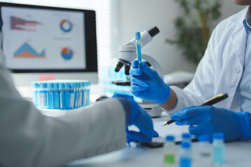 Medical researchers wearing lab coats and gloves are examining a test tube containing blue liquid,...